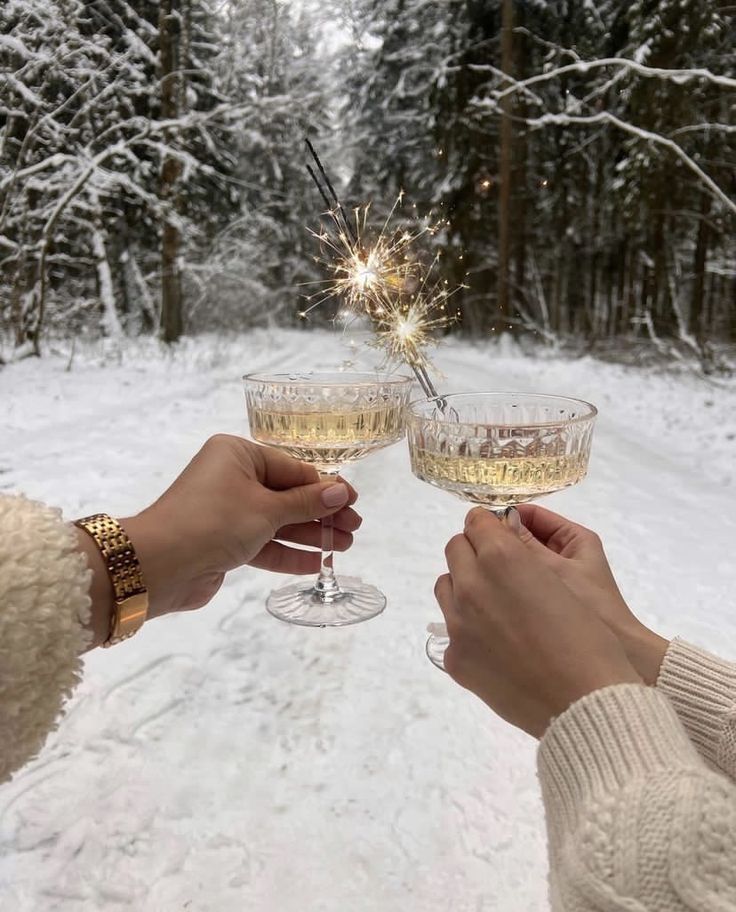 Two hands holding champagne glasses with a sparkler in a snowy forest setting.