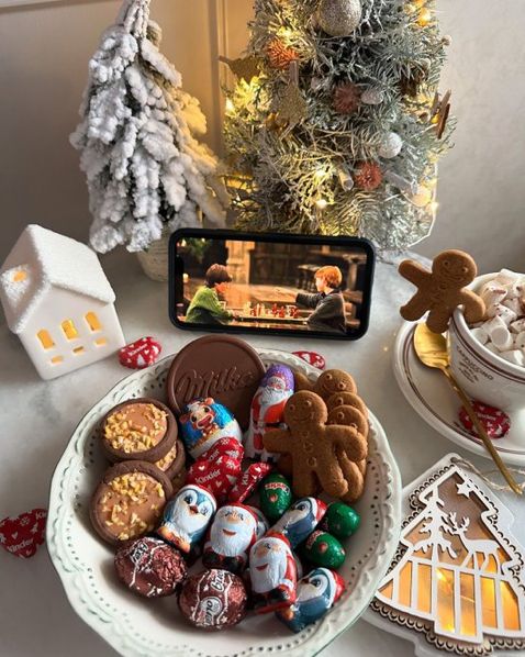 Festive treats and hot cocoa by a Christmas tree, with a cozy movie scene playing on a smartphone in the background.