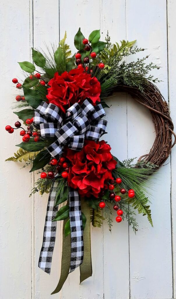 Festive wreath with red flowers, berries, greenery, and a black-and-white checkered ribbon on a white wooden background.