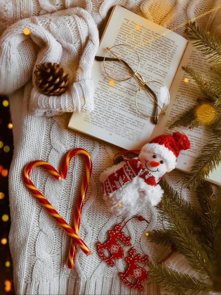 Cozy winter scene with open book, glasses, candy canes, snowman plush, pine branches, and sweater for Christmas vibes.