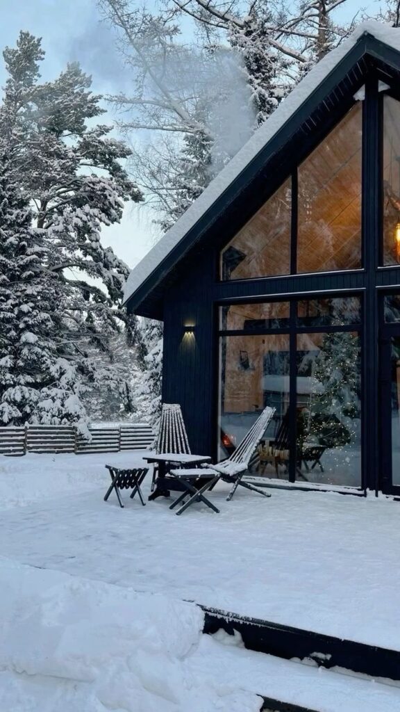 Cozy winter cabin with snowy landscape, large windows, and outdoor chairs, ideal for a peaceful retreat.