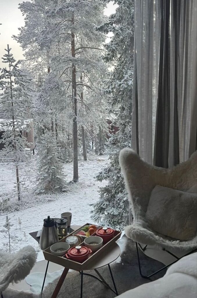 Cozy winter breakfast setting with a snowy forest view through a window, featuring tea and snacks on a tray.