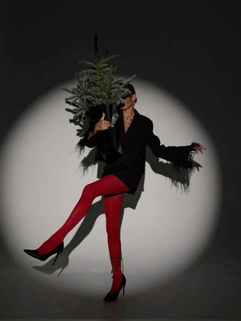 Fashionable person in red tights, black blazer, holding a small tree, posed dramatically in spotlight.