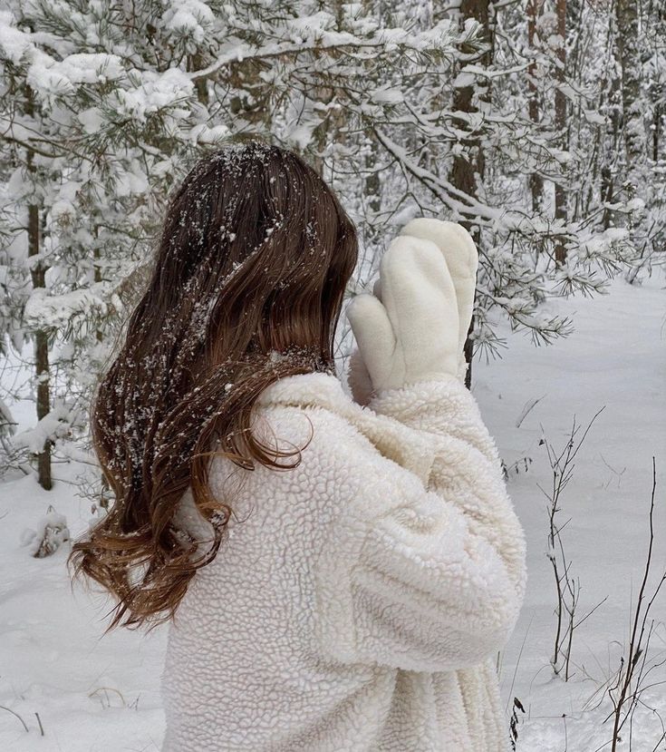 Woman in cozy white coat and mittens in snowy forest, with snowflakes on hair. Winter fashion and nature scene.