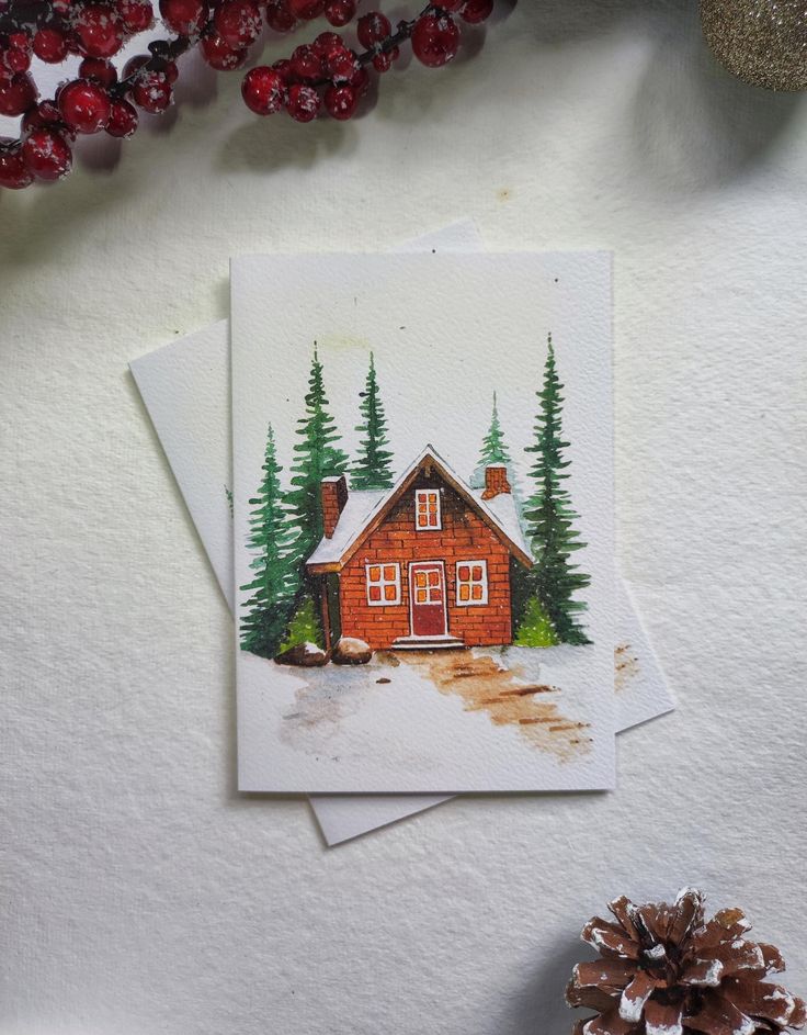 Watercolor holiday card with a cozy cabin and pine trees, surrounded by festive berries and a pinecone.