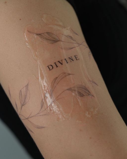 Fresh tattoo on arm with DIVINE text and delicate leaf design, covered in protective film.