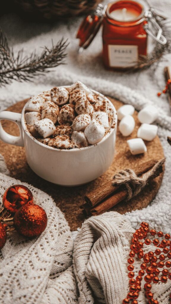 Cozy winter scene with hot chocolate topped with marshmallows, cinnamon sticks, and festive decor on knit fabric.