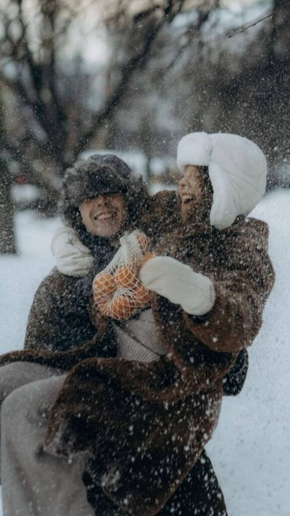 Couple enjoying a snowy day outdoors, bundled up in warm coats and hats, holding oranges.