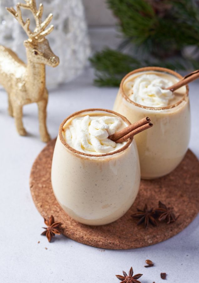 Festive eggnog with cinnamon sticks and whipped cream next to a decorative reindeer, perfect for holidays.