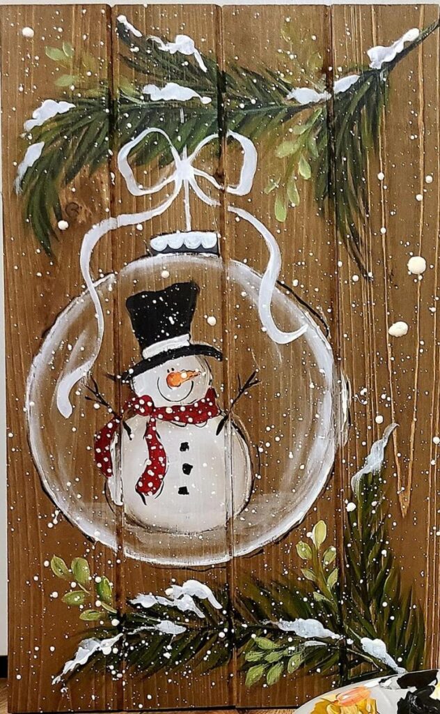 Snowman painted on wood inside a Christmas ornament with green branches and snowflakes.