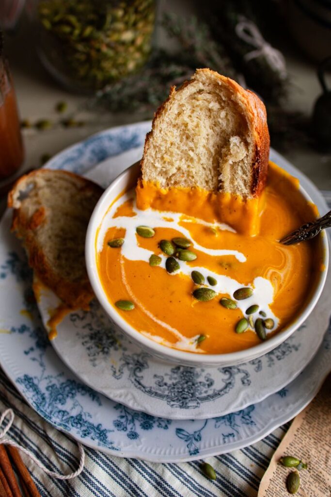 Creamy pumpkin soup topped with seeds, swirled with cream, and served with rustic bread on a floral plate.