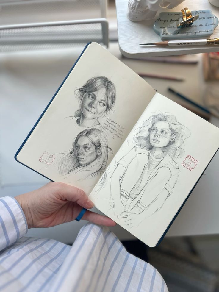 Hand holding sketchbook with pencil drawings of people, creative art tools on desk in background.