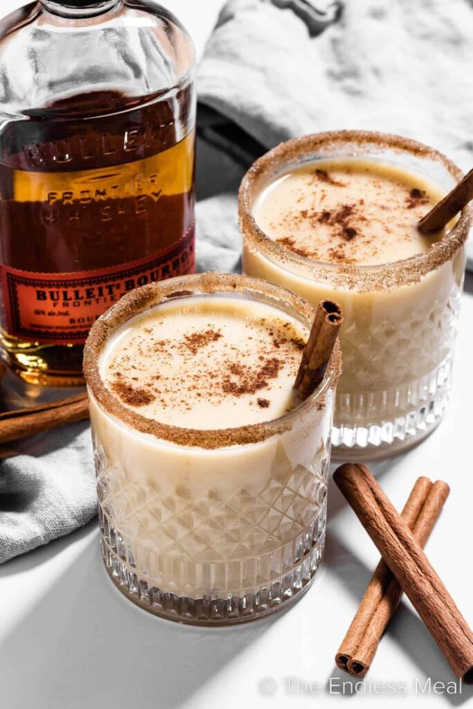 Two bourbon eggnog cocktails with cinnamon sticks, garnished with cinnamon sugar rims alongside a bottle of Bulleit bourbon.