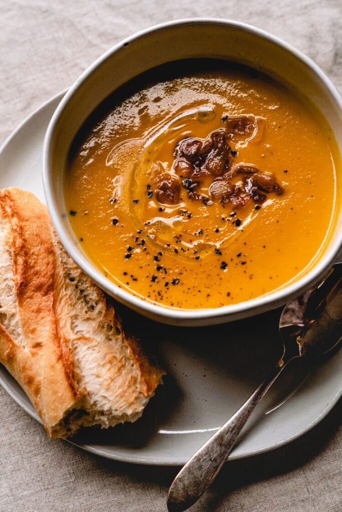 Creamy pumpkin soup with black pepper and a side of crusty bread on a plate, perfect for a cozy meal.