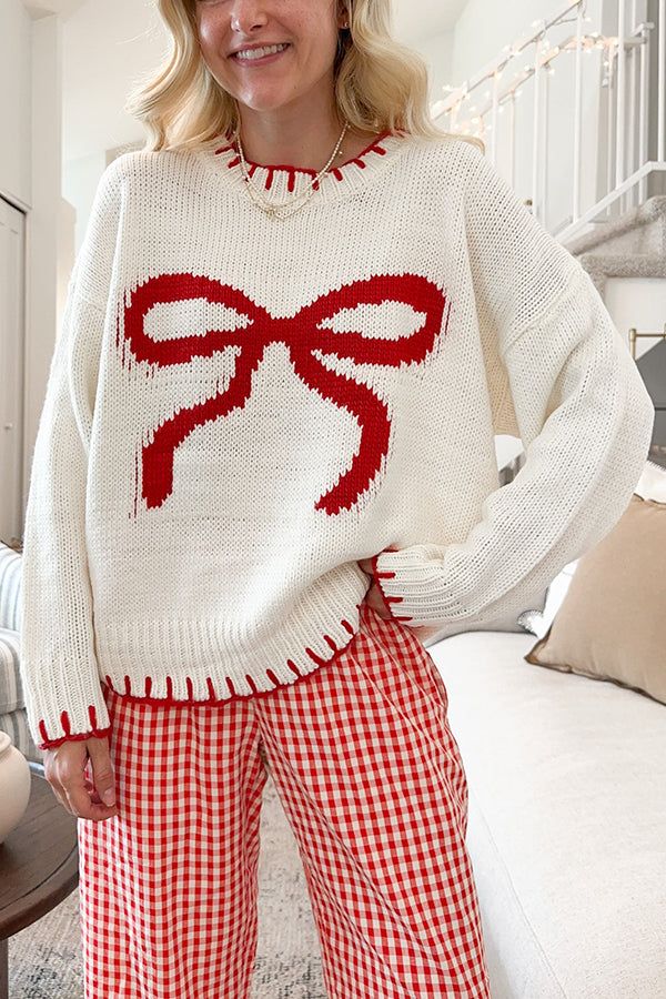 Woman in a cozy white sweater with red bow design and red checkered pants, smiling in a bright living room.