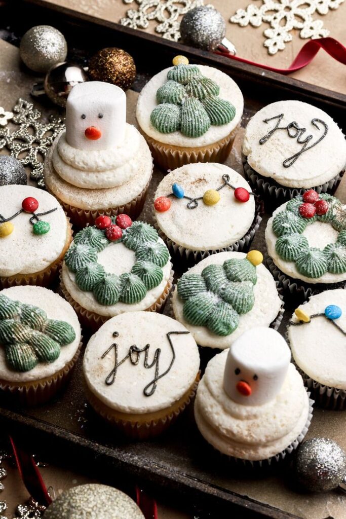 Festive cupcakes with holiday decorations, featuring snowman, trees, wreaths, and joy in frosting, surrounded by ornaments.