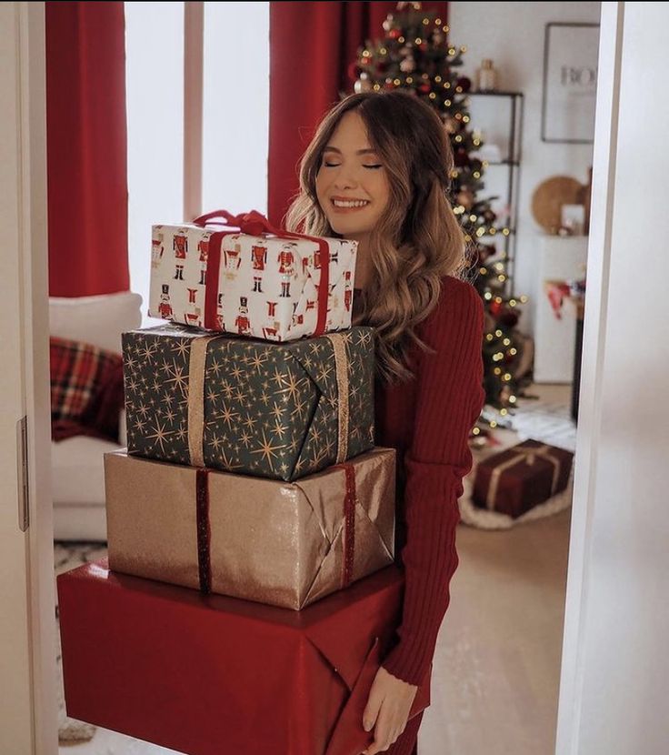 Woman in a cozy room holding stacked holiday gifts, Christmas decor in the background.