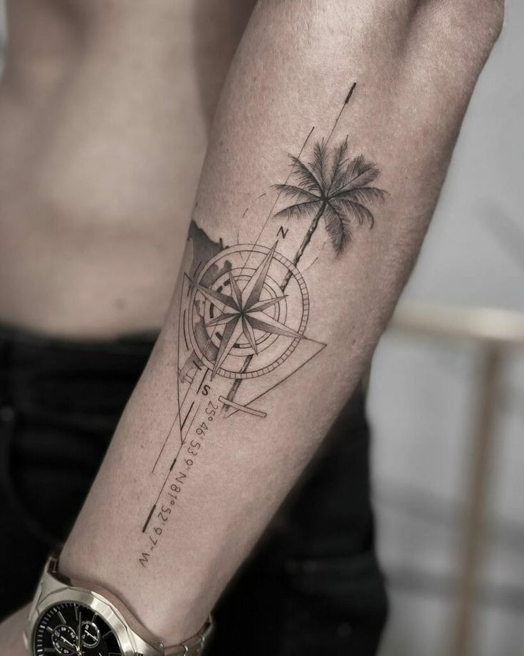 Forearm tattoo featuring a compass and palm tree design with geographic coordinates, worn by a person with a wristwatch.