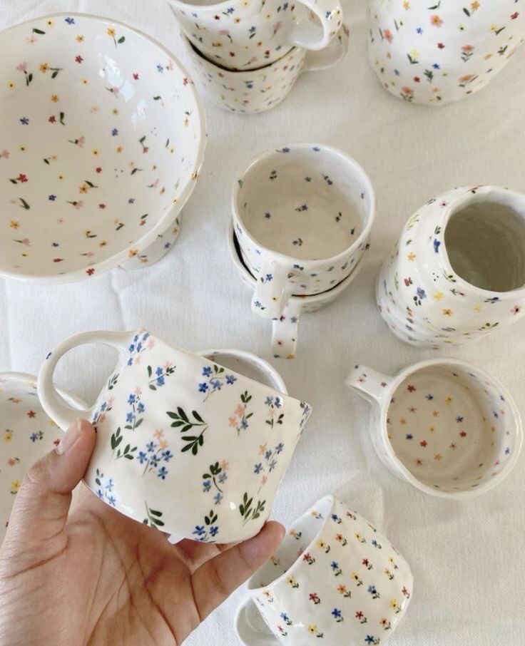 Hand holding a floral ceramic mug, surrounded by matching cups and bowls on a white tablecloth.