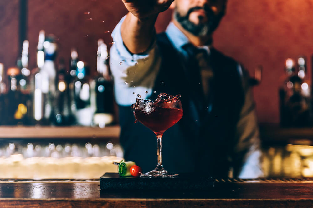 Bartender creating a splash with a red cocktail garnish in a dimly lit bar setting.