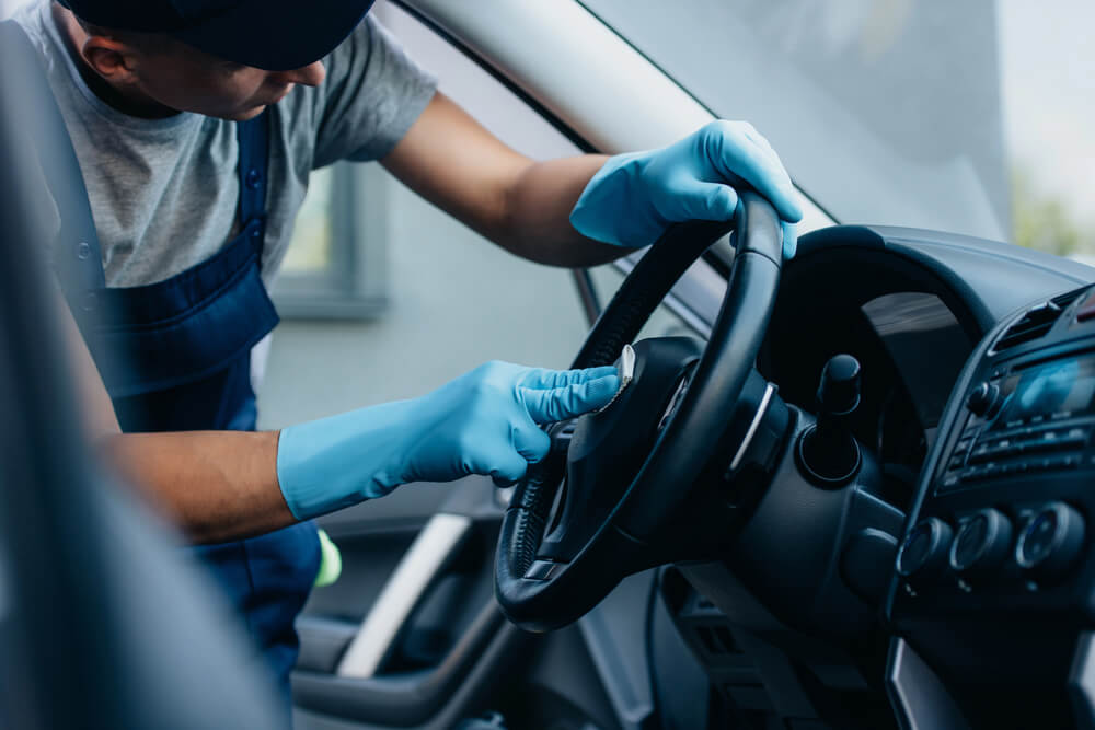 Worker cleaning car interior with gloves, focusing on steering wheel detail.