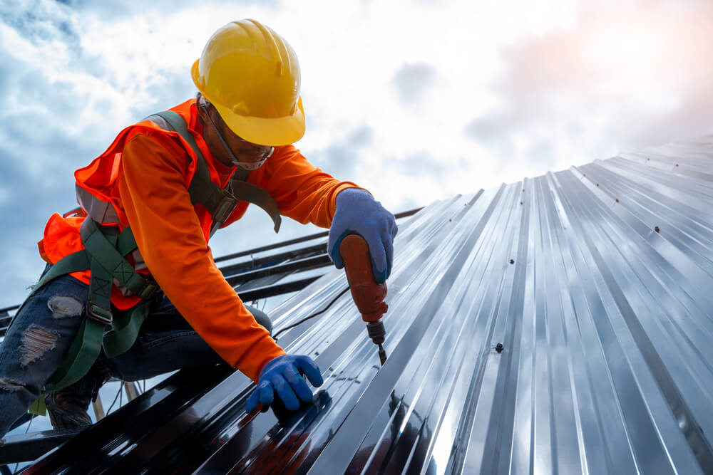 Construction worker installing metal roof sheet with drill, wearing safety gear under cloudy sky.