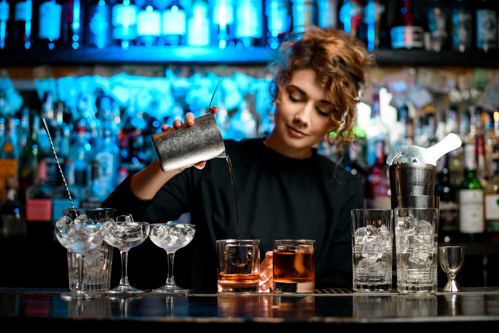 Bartender skillfully pouring cocktails in a dimly lit bar with various glassware and bottles in the background.