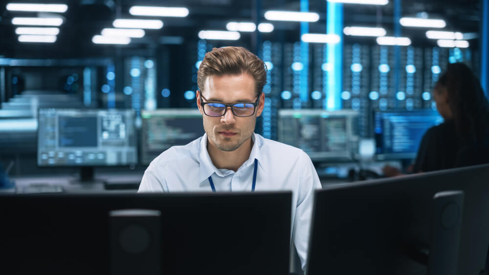 Person working on computer in server room with multiple monitors and digital data displays.