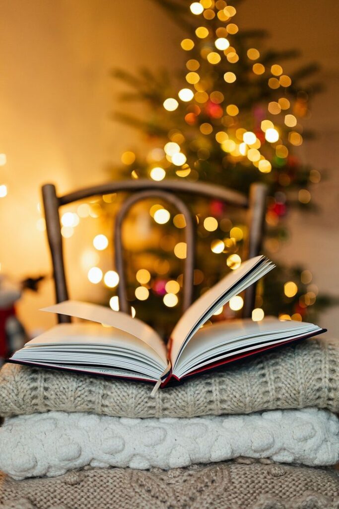 Open book on cozy knit blanket with Christmas lights and tree background. Holiday warmth and reading ambiance.