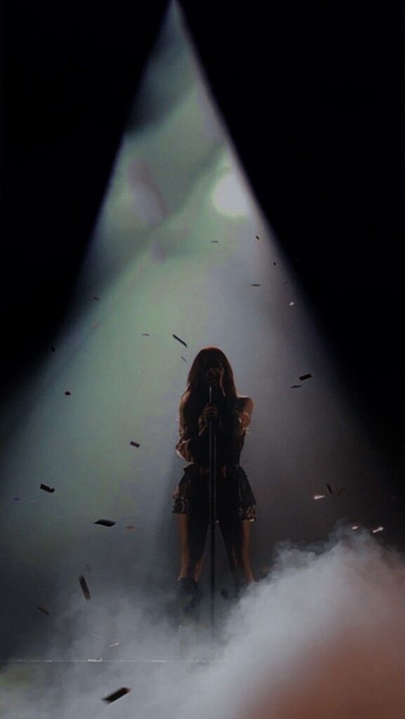 Singer silhouetted on stage performing under a spotlight with smoke effects and falling confetti.