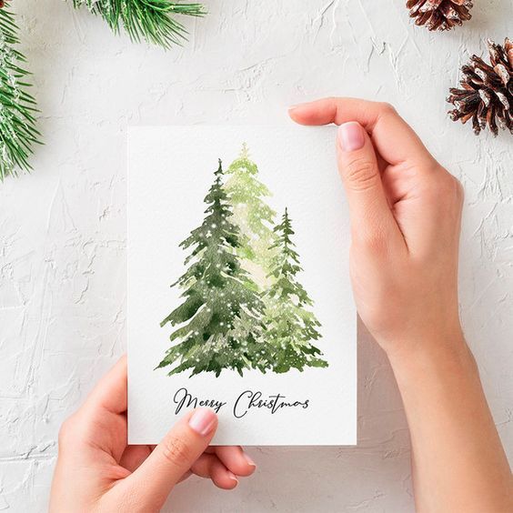 Hands holding a festive Christmas card with watercolor trees and Merry Christmas message on a textured background.