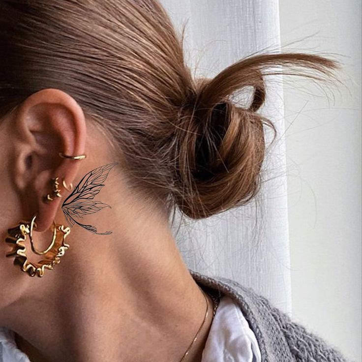 Woman with leaf tattoo behind ear, gold earrings, and messy bun hairstyle against white curtain background.