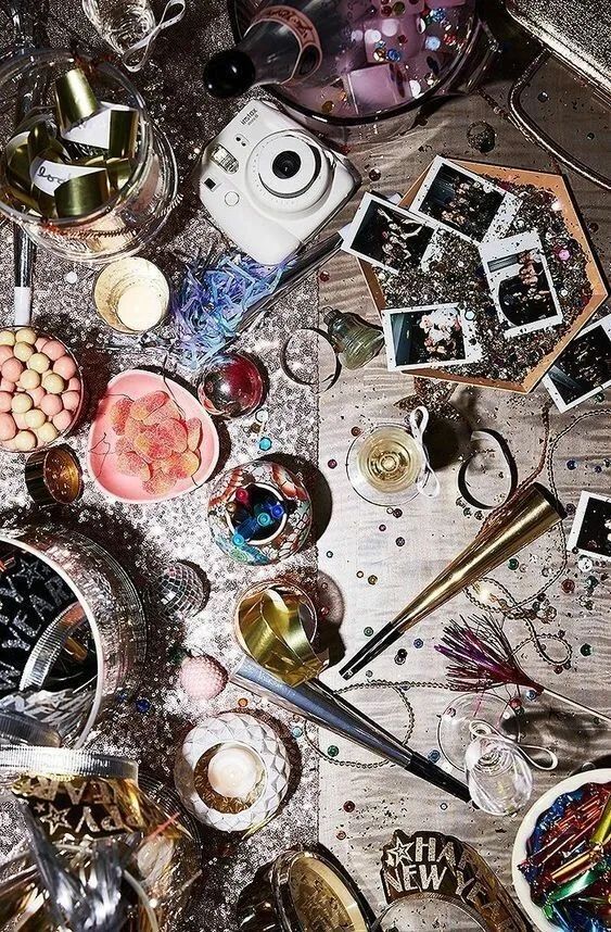 Festive New Year's celebration scene with confetti, drinks, instant photos, and party decorations on a glitter-covered table.