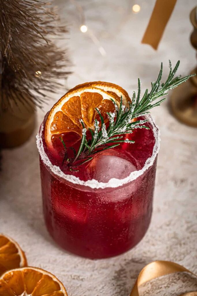 Festive red cocktail with rosemary garnish and orange slice in a salted-rim glass on a textured surface.