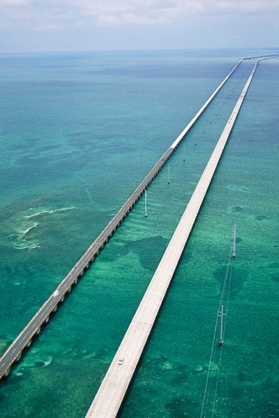 Aerial view of long bridge over turquoise sea, connecting islands under cloudy sky.