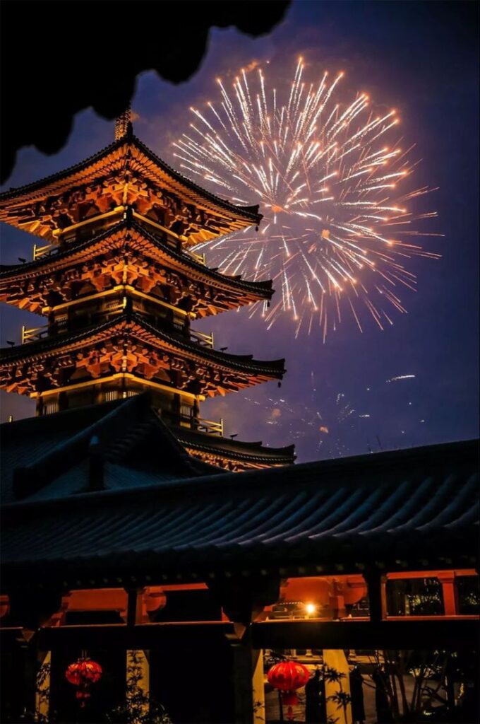 Illuminated pagoda with fireworks in night sky, celebrating festival with lanterns in traditional architectural setting.