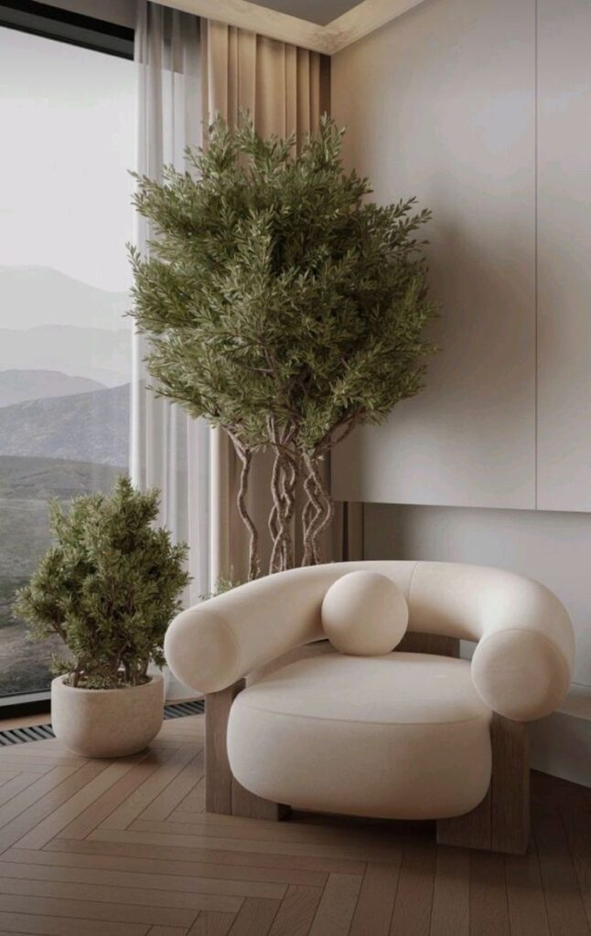 Modern living room with cozy beige chair, large potted tree, and serene mountain view through window.