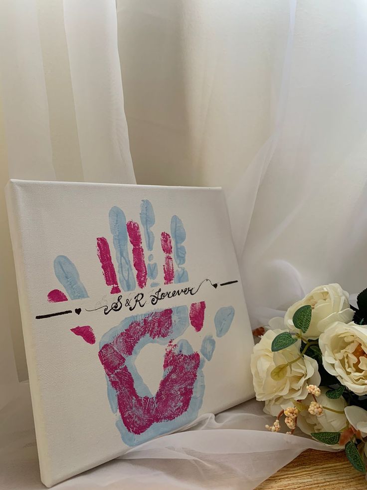 Handprint art canvas with S&R Forever message, surrounded by white flowers on wooden table, soft curtain backdrop.