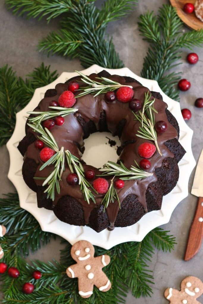 Festive chocolate bundt cake with rosemary, raspberries, and cranberries on a white plate surrounded by holiday decor.