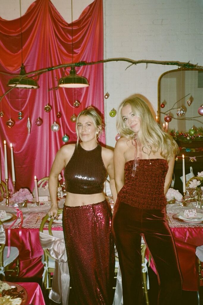 Two women in festive outfits pose at a decorated party table with pink and red holiday ornaments and drapery.