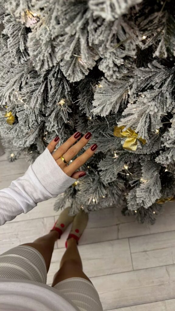 Person with red nails touching a decorated snowy Christmas tree with white lights and gold ribbon.