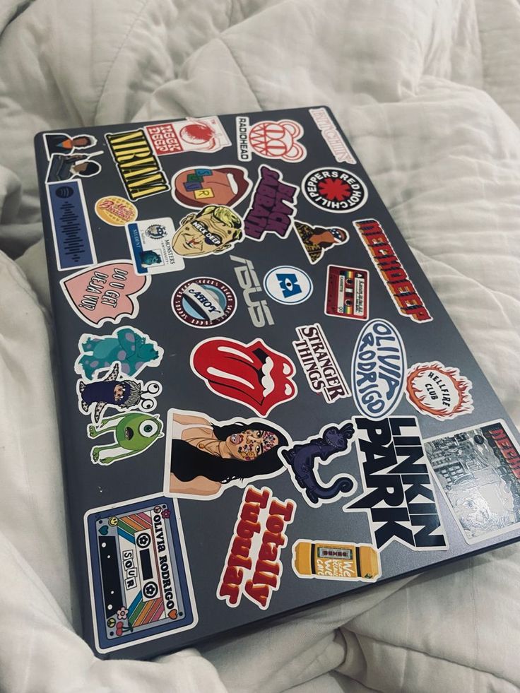 Laptop with diverse stickers featuring bands, retro designs, and pop culture icons, all on a cozy bed setting.