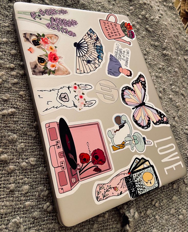 Laptop with diverse aesthetic stickers, including floral, animal, and motivational designs, on a textured surface.