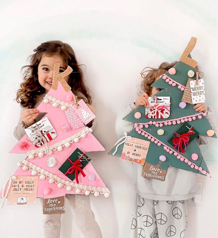 Children holding handmade holiday gift card displays shaped like Christmas trees, decorated with tags and pom-poms.