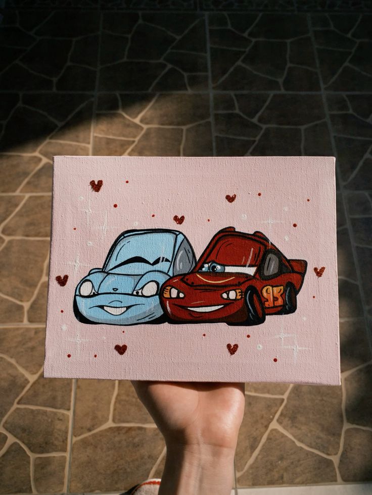 Cute hand-painted car characters on canvas with hearts, held against a stone tile floor background.