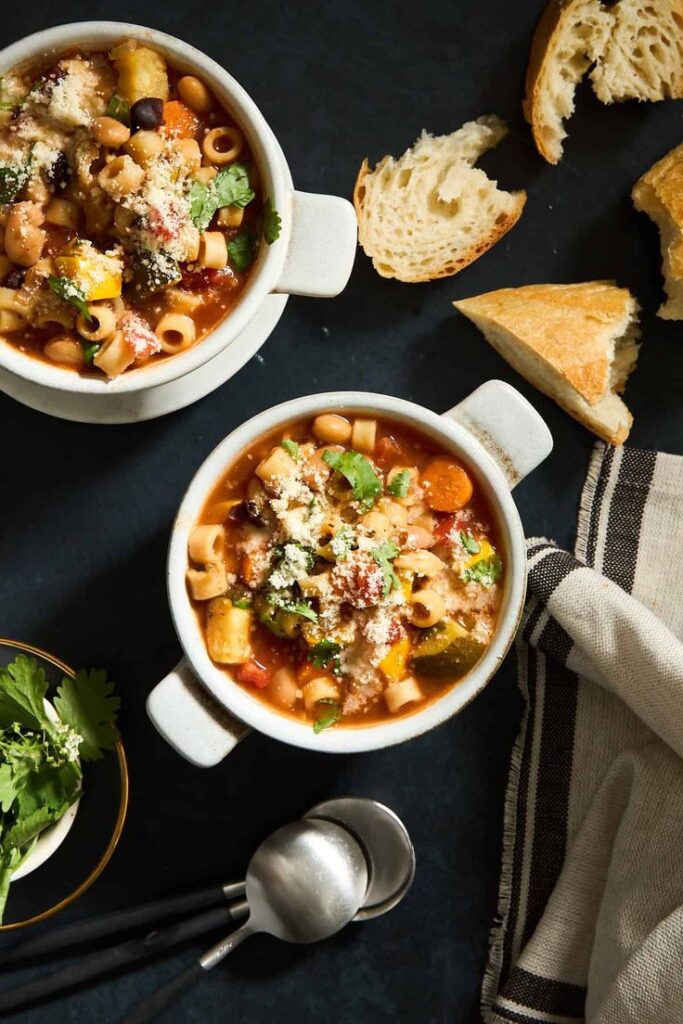 Two bowls of hearty minestrone soup with pasta and vegetables, served with rustic bread on a dark table.