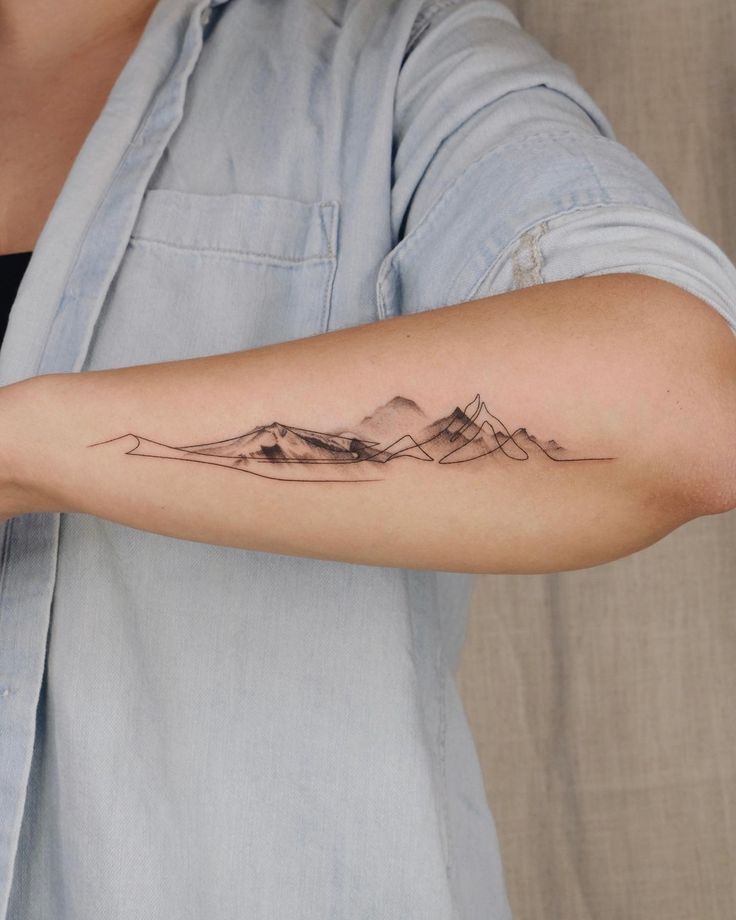 Minimalist mountain tattoo on forearm, person wearing denim shirt.