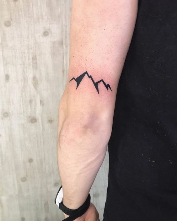 Minimalist mountain tattoo on inner arm with a wooden background.