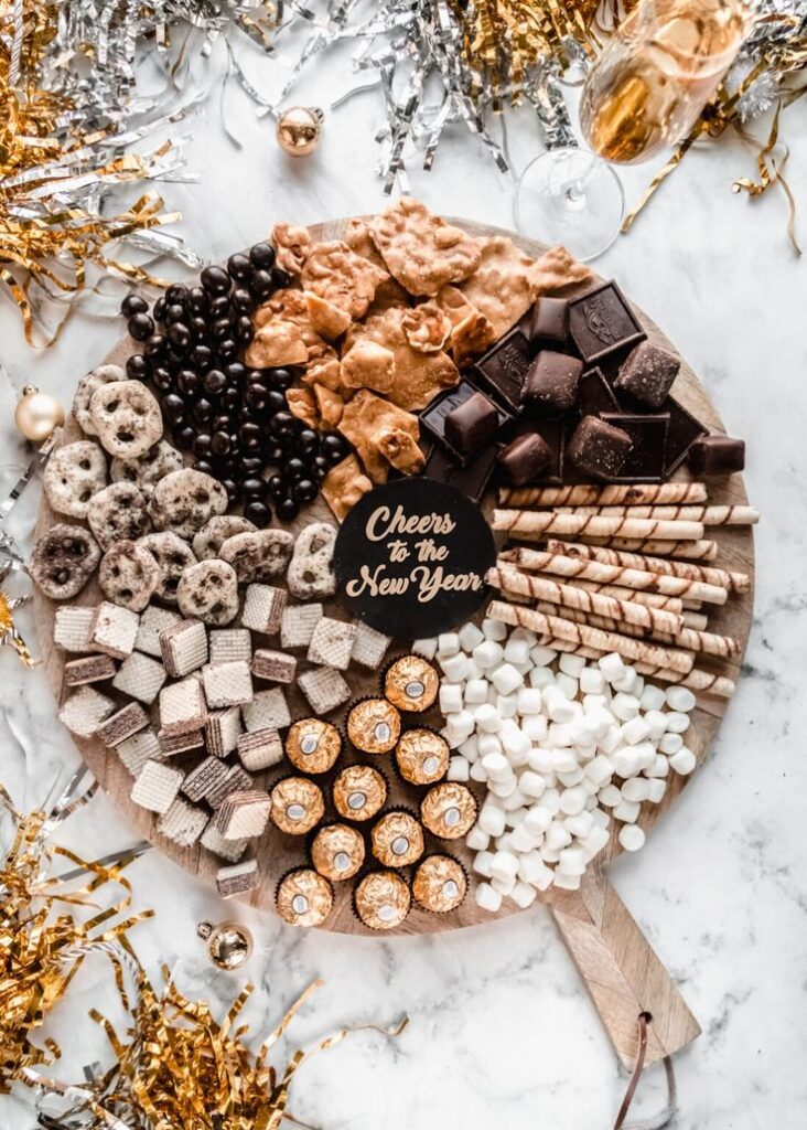 Festive New Year dessert platter with chocolates, wafers, and marshmallows on a marble table decorated with confetti.