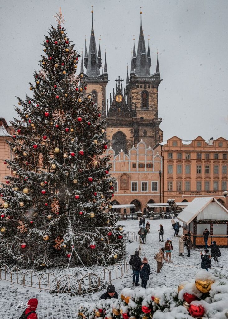 Snowy Christmas market scene with a decorated tree and historic church in Prague's Old Town Square.